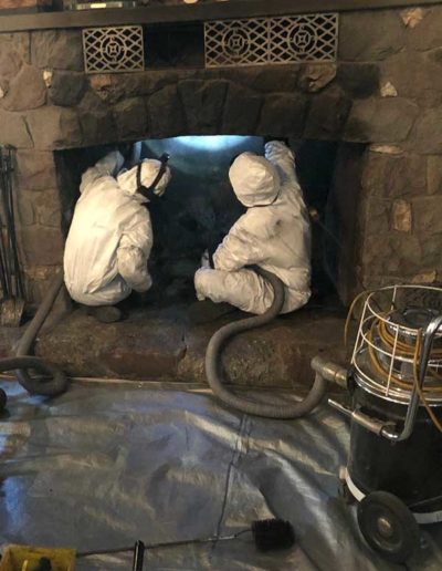 Two men in white cleaning suits sitting in fireplace using vaccums and a grey drop cloth used in front