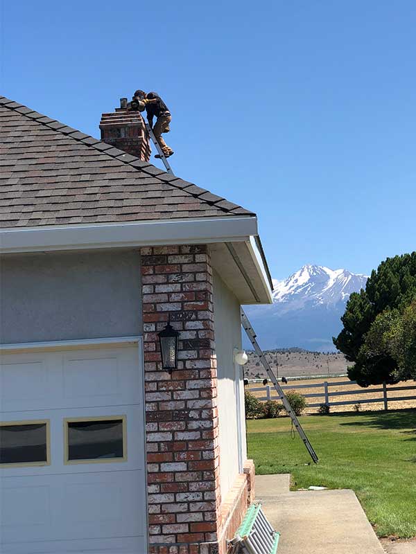 man on a ladder working on chimney of home mountains are seen in the background