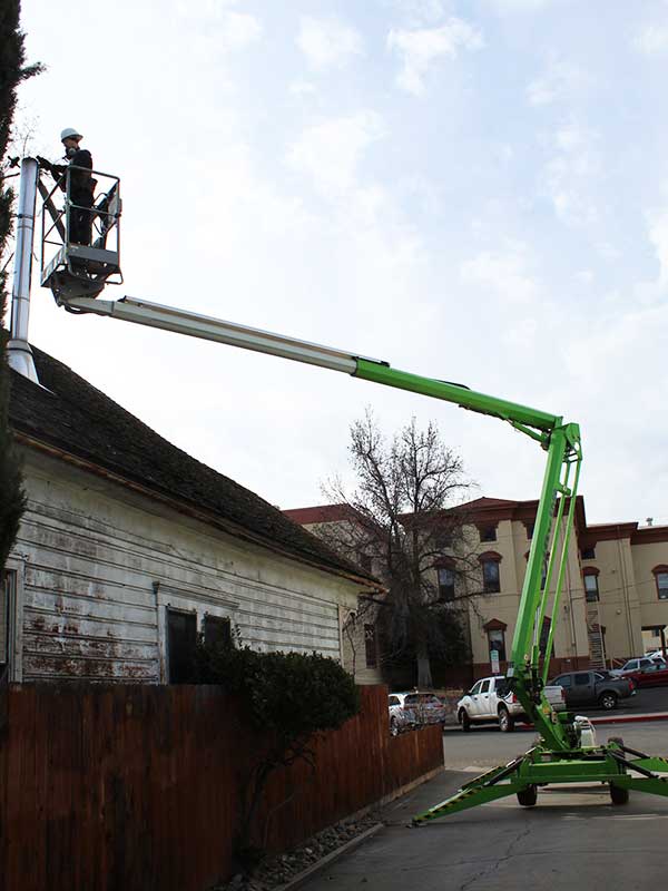 man on the bucket of a lift is raised above a roof of a home to inspect the chimney