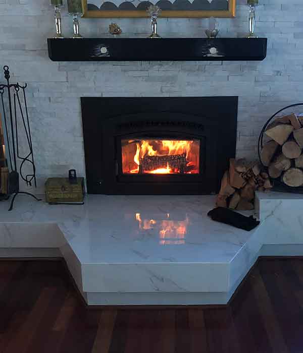 Fire burning in black fireplace insert white tile and hearth around it with chimney tools and extra firewood on the sides 