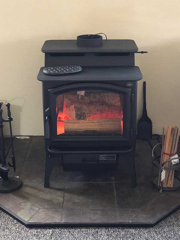 Black wood stove with fire burning in it with wood next to it on a tile floor piece