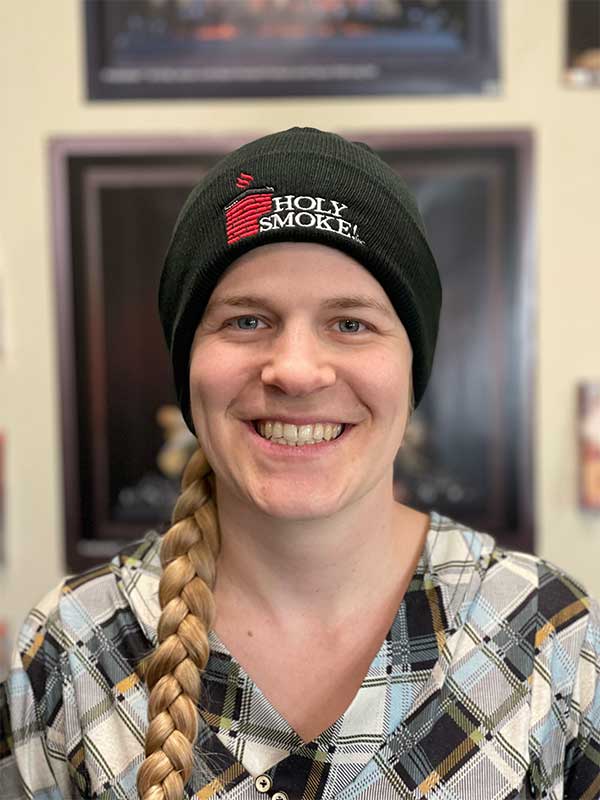 Woman wearing black sock hat that says Holy Smoke hair is in a braid and wearing plaid shirt
