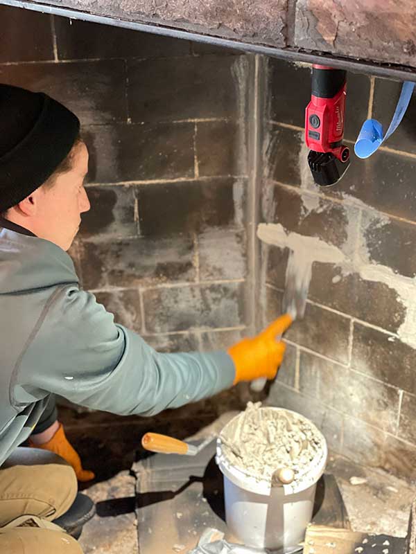 Man in grey hoodie and black sock hat has a bucket of material that he is applying and repairing the maosnry on the firebox of a chimney