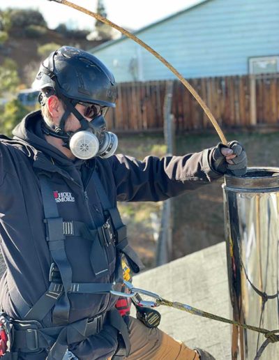 man wearing black helmet and jacket with respirator using cleaning tool down chimney