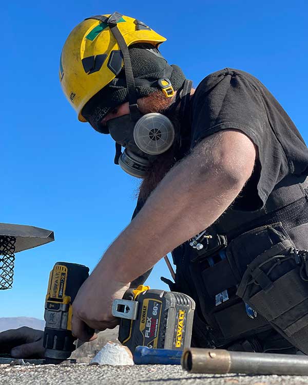 man wearing yellow helmet and breathing mask holding a drill and working on tile of a chimney on a roof